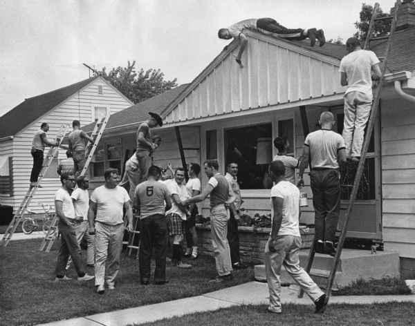 Several men, many wearing T-shirts, are gathered on the lawn outside a house, with some men standing on ladders. One man is lying on the roof and painting the side of the house. Caption reads: "<b>POLICEMEN PAINT INJURED OFFICER'S HOME</b> — Off duty Milwaukee policemen swarmed over a house belonging to Patrolman Ralph Waters Thursday as they applied a coat of primer. They will paint the house Sunday. Walters, of 3134 S. 55th st., suffered a skull fracture in the recent rioting."