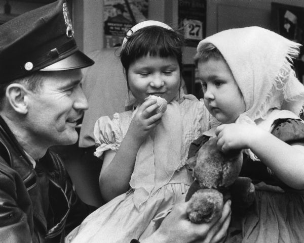 A uniformed police officer is handing a teddy bear to a young child, as another child is eating and smiling. Caption reads: "<b>Wauwatosa's police station</b> was host to a couple of wandering little girls for several hours Thursday. Patrolman Ray L. Rutowski obtained ice cream for Frances Figuaroa (left), 4, and her sister, Mitzi, 3, daughters of Mrs. Charlotte Figuaroa, a divorcee, of 2424 N. Humboldt av. Wauwatosa police took the girls off a Transport Co. bus at Milwaukee av. and Church st. after the bus driver realized that they were riding without adult supervision. They apparently had started joy riding on buses shortly after their mother had let them out to play early Thursday. The children were turned over to a grandmother while a conference between the mother and children's court authorities was being arranged. The mother said she had not missed the children because she was busy cleaning house."