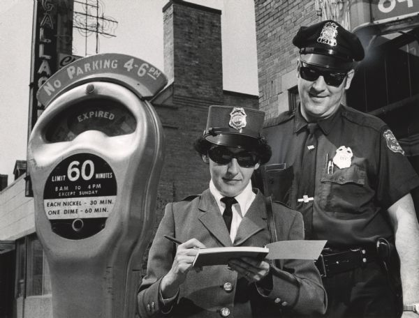 A uniformed female officer writing in a book, as a uniformed male officer is smiling and looking on over her shoulder. A parking meter is in the foreground. Caption reads: "THE MILWAUKEE POLICE Department put its first woman meter maid on the job this week. Mrs. Ruth Niedfeldt wrote a traffic ticket under the watchful eye of a regular traffic patrolman, Glenn Wichgers, who accompanied her on her first rounds. She covers the area bounded by River St. to N. 3rd St., Kilbourn Ave. to Michigan St."