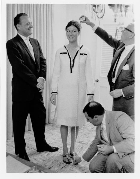 Actress Gia Scala smiles as two men measure her height with a tape measure. Movieland Wax Museum founder and president Allen Parkinson stands next to her and smiles. She is being measured for a wax figure at the museum.