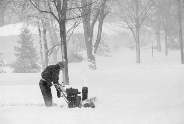 A man is using a snowblower during a blizzard. A tree-lined street with a row of houses is in the background.