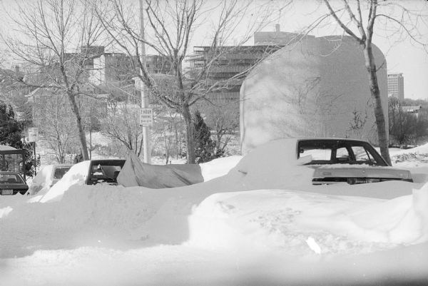 View down Gorham Street towards cars covered in snowdrifts. James Madison Park and Mansion Hill are in the background.