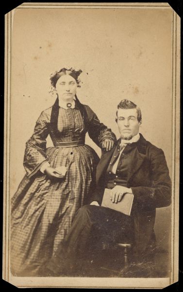 Carte-de-visite of John and Martha (Sjurson) Anderson on their wedding day. Martha is standing and is resting her hand on John's shoulder. John is sitting in a chair holding a book in his lap.