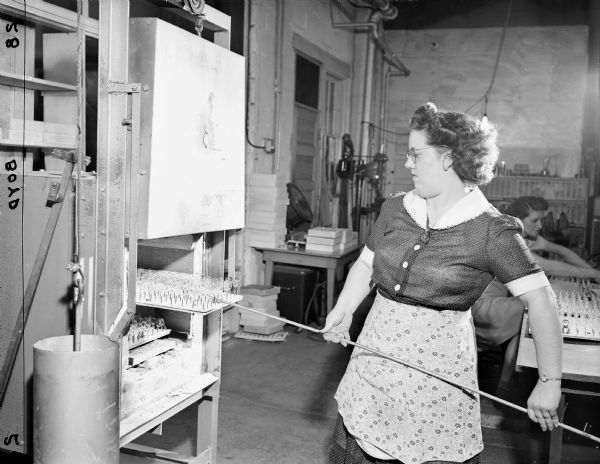 A woman is using a metal rod to handle glass objects on a tray. Caption reads: "Women workers at Chaney Instruments." Another woman in the background is working with another tray of the glass objects on a low table.