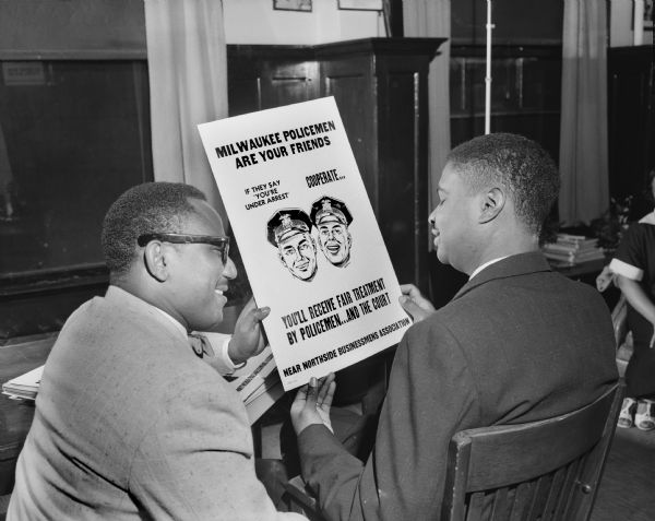 Two African American men smiling while holding a poster. The poster includes a drawing of two smiling police officers and the text: "Milwaukee Policemen are your Friends. If they say 'You're under arrest' Cooperate... You'll receive fair treatment by Policemen...and the Court. Near Northside Businessmens Association." There is a woman sitting in the background on the right.