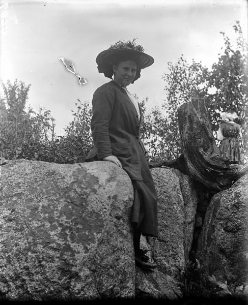 Portrait of a woman wearing a hat sitting on rocks near a tree stump that has roots growing down between the rocks. Another woman is standing just behind the stump in the background on the right, with her back turned away from the camera. Caption identifies the location as Big Falls.