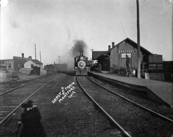 A locomotive is moving along the tracks along the platform at the depot in Montfort. A man is standing near carts on the right at the end of the platform, and a group of people are standing on the platform in front of the depot. The photographer's shadow is in the foreground.