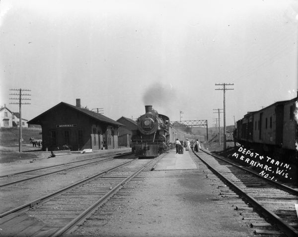The Merrimac depot and a train. A group of people are standing in between the train tracks near the railroad cars.