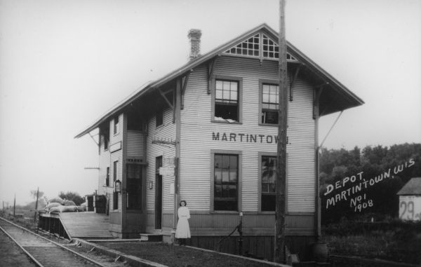 Exterior view of the Martintown train depot. A woman or girl is standing in front of the building. Caption reads: "Depot, Martintown, Wis."