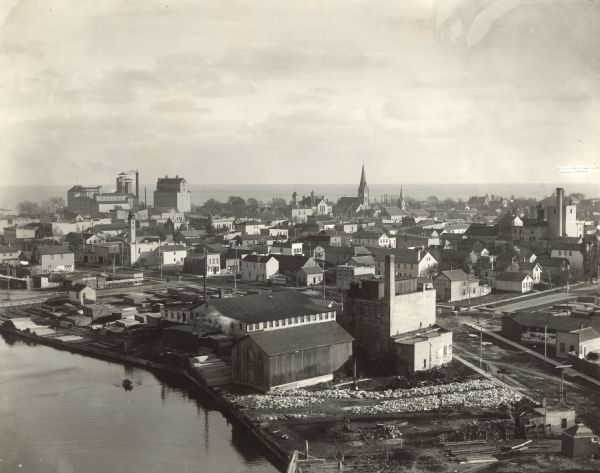 Bird's-eye view of Manitowoc taken from Northern Grain Company's Elevator B, facing southeast. South Main (Tenth) Street intersects with Franklin Street in center-left. Along the city skyline, the William Rahr Sons brewery, malt house and elevator complex is on the left. The First German Evangelical Lutheran Church and School are in the center, with Lake Michigan in the background. In the foreground, along the Manitowoc River from left to right are Chris Schoch Lumber Yard, Bigel & Guse Planing Mill & Building Contractors and the mill and warehouse previously known as Wisconsin Central Mills, owned by Jacob Fliegler. The three-story brick mill has a dust collector room on the roof. A spur track crosses South Water Street. In 1892, fire destroyed the Manitowoc Manufacturing Company, which occupied the triangle-shaped block between Franklin, South Twelfth and South Water Streets. The triangular corner of the block is in view on the right.  The fire resulted in the failure of the T.C. Shove Banking Company and consequent closing of Jacob Fliegler's and Truman and Cooper's mills.  The bank had been heavily invested in all three concerns, most heavily in the Manitowoc Manufacturing Company, and the huge fire loss caused it to be insolvent. Fliegler's mill was unoccupied for several years as lawsuits were settled and property sold. In 1898, the Manitowoc Pea Packing Company was formed and factory buildings erected on the site of the 1892 fire. Fliegler's mill, across the street, was purchased in 1899 and remodeled for its use as a warehouse, cold storage facility and offices.
