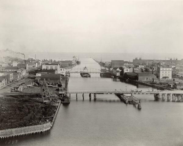 Bird's-eye view of the Manitowoc Harbor facing east from Northern Grain Company's Elevator B. In the foreground, Mathews & Keith's pile driver scow is secured along the dock line on the left. Spanning the Manitowoc River are the Main (Tenth) Street and Eighth Street swing bridges. At the mouth of the river, a ship is moored at the Goodrich Transit Company docks. Opposite the docks, the entrance to the Chicago and Northwestern Rail Road's new car ferry slip is partially visible. A long coal shed with four "travelers" on the roof is to the right. In 1892, Congress granted approval to the U.S. Army Corps of Engineers for construction of a ship channel, twenty feet in depth, connecting the waters of the Great Lakes between Chicago, Duluth and Buffalo for the improvement of commerce on the lakes. All sections of the deepened channel were to be completed in 1895. Manitowoc, with a river depth of fourteen feet, began a massive program of harbor improvements in 1895 in order to accommodate expected larger vessels with twenty-foot drafts. At the same time, the Wisconsin Central Rail Road was constructing a rail line into the city with car ferry service to Michigan and making harbor improvements, as well. In June of 1898, dredging between the U.S. government piers at the river's entrance was completed. The channel, up to the new turning basin adjacent to Burger and Burger Boat Yard, had attained a depth of twenty feet.