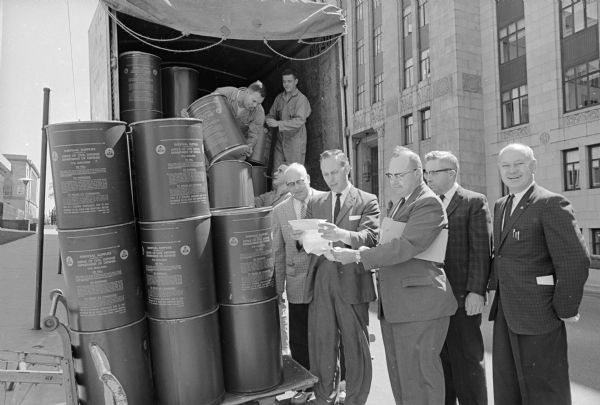 Barrels of supplies to stock area fallout shelters being unloaded at the City-County Building by work relief program participants and Huber law prisoners from the Dane County jail. Checking in the supplies at right are: City Civil Defense Director, Richard C. Wilson; Dane County Civil Defense Director Curtiss Brauhm; and Wilson's assistant, Mello Stapleton.