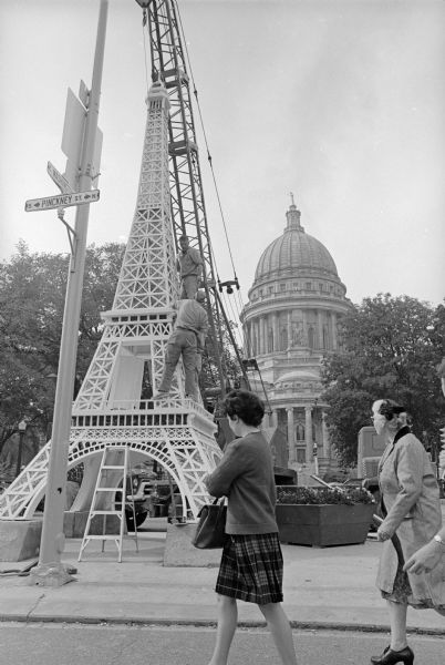 Pedestrians pass a replica of the Eiffel Tower being constructed by workers at the corner of East Washington and Pinckney Streets on the Capitol Square. The Wisconsin State Capitol is in the background. The replica was installed for Festival de France which took place in Madison from September 27-October 3, 1964.