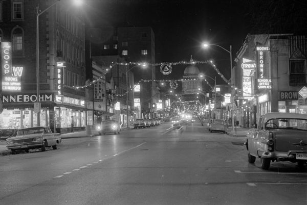 Night view of State Street taken from Lake Street intersection and showing Christmas season lighting. A number of businesses are seen, including Rennebohm's, Brown's Books, Troia Steak House, and Warner Medlin Photographers. Cars are parked on both sides of the street. The Wisconsin State Capitol building is at the opposite end of State Street.