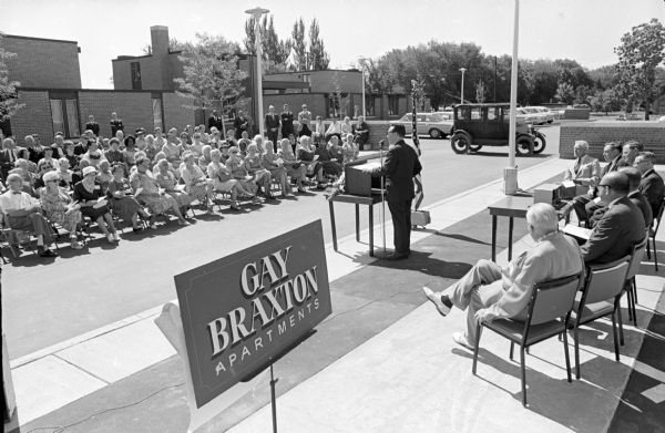 Roland Day, at the microphone, emcee of the dedication of the Gay Braxton Apartments. Speakers seated behind Mr. Day include Mayor Otto Festge and tenant spokesman, Fred Wildeman. A large group of spectators sitting in folding chairs are listening to the speaker. A vintage Ford owned by tenant Alphonzi Chiovaro is parked nearby.