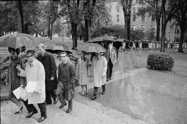 A long, orderly line of student sorority and fraternity members carrying umbrellas and marching in pairs to protest the decision to ban the Delta Gamma sorority from the UW-Madison campus. They are walking up the stairs to Bascom Hall. Protestors walked up "Fraternity Row" on Langdon Street, up Bascom Hill, past Bascom Hall where they were met by Dean of Students, LeRoy Luberg, who was presented with a protest resolution by Robert Jennings, President of the Wisconsin Intrafraternity Association. The group of about 2,000 then walked back down the hill and back to Langdon Street. The decision to ban the sorority came after the national sorority suspended the Beloit chapter for pledging Patricia Hamilton, an African-American woman from Madison. The group is seen here nearing Bascom Hall with North Hall in the background.