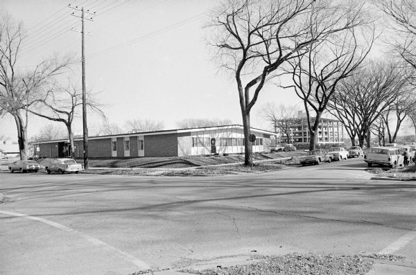 View of the new Neighborhood House at 29 S. Mills Street taken from the intersection of S. Mills and Milton Streets.   It was dedicated Oct. 31, 1965. The building housed both the Neighborhood House and the West Clinic for the Dane County Mental Health Center. Several cars are parked on the street, and the Meriter Clinic at 20 S. Park Street is under construction in the background.