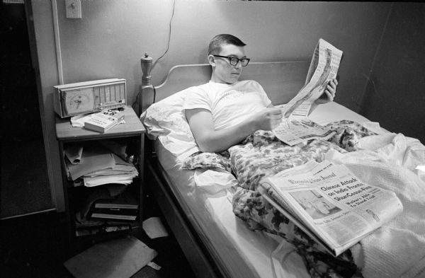 U.W. student-athlete Pat Richter relaxing at his home at 2034 Yahara Place before his final home game for the Badger football team. He is lounging in bed reading the newspaper, with a radio and books on a nightstand nearby.