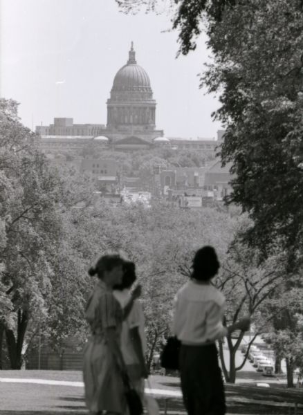 View of the Wisconsin State Capitol from Bascom Hill, with three women walking in the foreground. Cars are parked on State Street at the base of the hill. Several State Street establishments, including the Capitol Theater, can be seen in the distance in front of the Capitol.