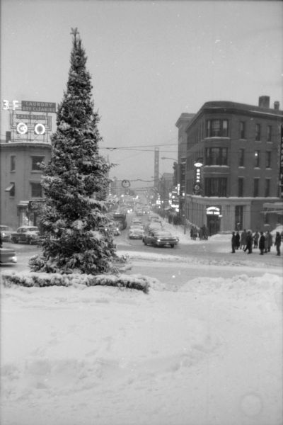 View down State Street from the Capitol Square following a snowstorm. A Christmas tree is standing on the corner, and other holiday decorations are hanging over the street. Several pedestrians are waiting to cross the street as traffic circulates along Mifflin and State Streets. Several State Street establishments can be seen including the Orpheum Theater and the Commercial State Bank.