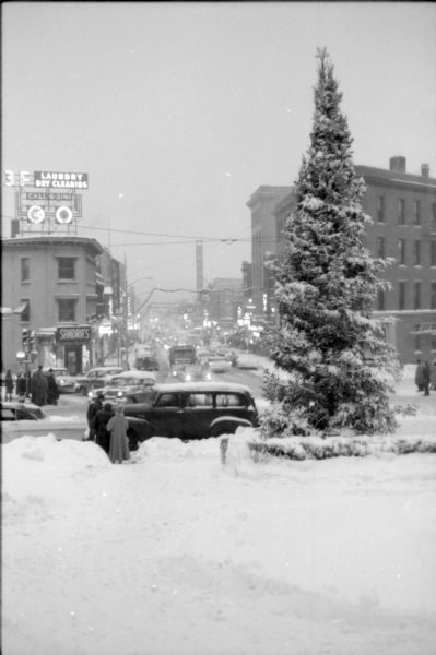 View down State Street from the Capitol Square following a snowstorm. A Christmas tree is standing on the corner and other holiday decorations are hanging over the street. Several pedestrians are waiting to cross the street as traffic circulates along Mifflin and State Streets. Several State Street establishments can be seen including the Orpheum Theater and Sandra's.