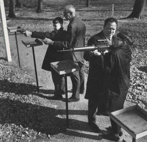 Two men assisting two children in firing guns. Caption reads: "Shooting on a rifle range is part of the program for the Sauk county junior deputy sheriff program. Undersheriff Ralph Hearn helped Mark Olson, 11, Reedsburg, fired [sic] a pistol and Deputy Mike Spencer aided Conrad J. Ligel, 11, Plain, shooting an AR-15, the police version of the M-16 rifle."