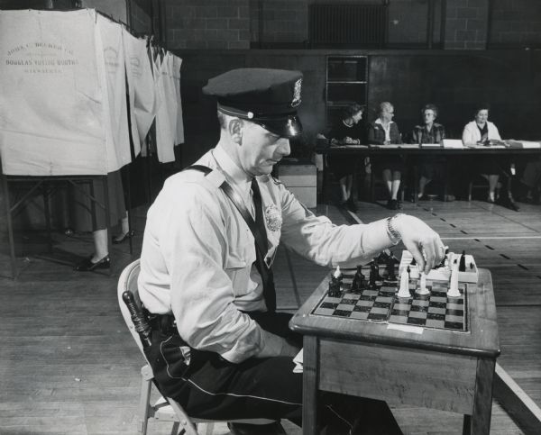 A uniformed police officer is sitting in a folding chair and moving pieces on a chessboard. Caption reads: "<b>On duty Tuesday</b> at the voting place in the basement of Riverside high school, 1615 E. Locust st., Patrolman Edward Evenson, of 2644 N. 17th st., relieved some of the monotony by playing a game of chess against himself."