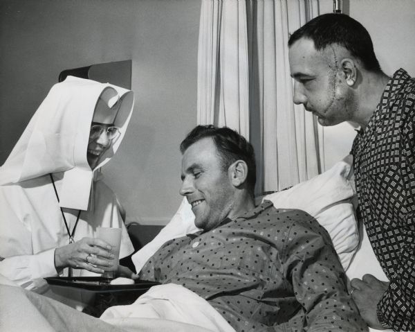 A nun is handing a glass to a man in a bed. Another man — who has numerous stitches along the side of his face — is standing next to the bed. Caption reads: "<b>Recovering from knife wounds,</b> two Milwaukee policemen share the same room at Misericordia hospital. The officers, William G. Kasten, 23, of 3625 N. 82nd st., and Kenneth G. Stanelle, 29, (right) of 1945 N. 28th st., were slashed in a knife-gun fight on Thanksgiving night. They talked with Sister Immaculate Heart of Mary, S.M."
