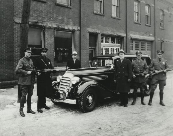 Four uniformed police officers holding guns are posing alongside a car, with two other men. A brick building behind them has a sign on the window that reads: "Department of Police." Caption reads: "1935 Buick Armored Car. From left: Carl Radtke; Alma E. Thomack (?); Bill Williamson — sold arms and ammunition; Police Chief George T. Prim; Al Deltgen; Fred Arndt. Thompson sub-machine guns (as noted on reverse of original). Original at Outagamie Co. Historical Society."
