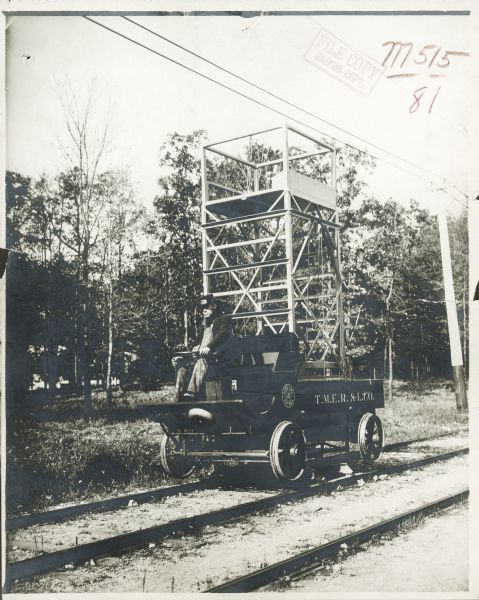 A man is driving a line car with scaffolding on the car. The signs on the side read: "Auto Line Patrol 10" and "T.M.E.R. & L Co." The car is set up on a platform so that the wheels are just above the railroad tracks. Power lines are overhead.