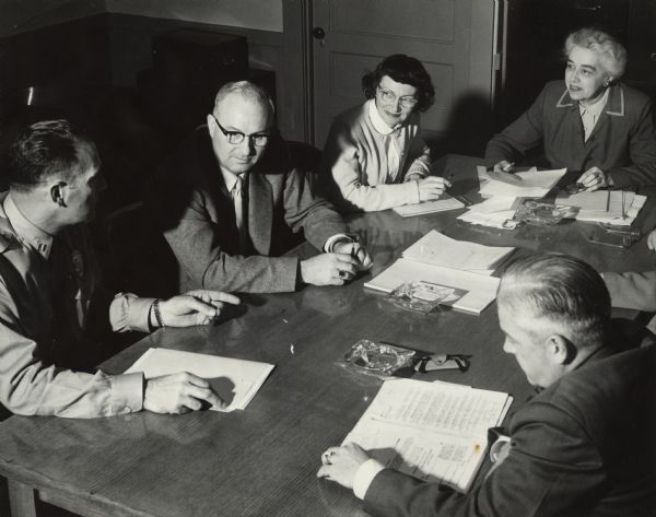 Slightly elevated view of several people sitting around a table with papers in front of them. Caption reads: "Meeting of City Police and Fire Commission. 7:30 p.m. Wednesday May 19, 1954 at the Central Fire Station (No. 1). Left to right: Captain Richard Gruber, 1013 East Gorham; Thomas J. Doran, 4117 Yuma Drive; Mrs. Helen W. Hevey, 3629 Sargent, stenographer for the commission; Mrs. Mary J. Tegge, 149 W. Wilson; and in the right foreground, Chief Edward J. Page, 429 West Johnson; J. 10 Assignment taken by: James K. Meyers; Instructor: Mrs. MacLean."