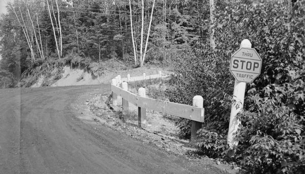 View of curve in unpaved road, with a guard rail on the right. There is erosion underneath trees just beyond the curve of the road. A road sign on the right reads: "STOP Thru Traffic." Trout Lake (vicinity). Caption reads: "'Very Accidental' intersection."