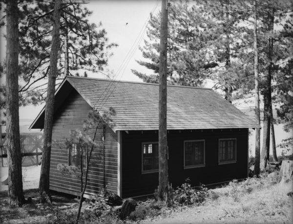 View looking down slope towards the Bacteriology Laboratory on the shore of Trout Lake. Trees are around the small, wood sided building, and the lake is in the background.