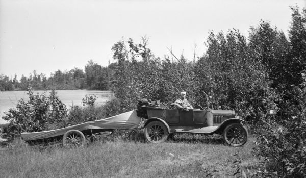 Caption reads: "Dr. Birge, probably at Trout Lake." He is sitting in a car. There is a boat covered by a tarp on a trailer behind the car. A lake is in the background.
