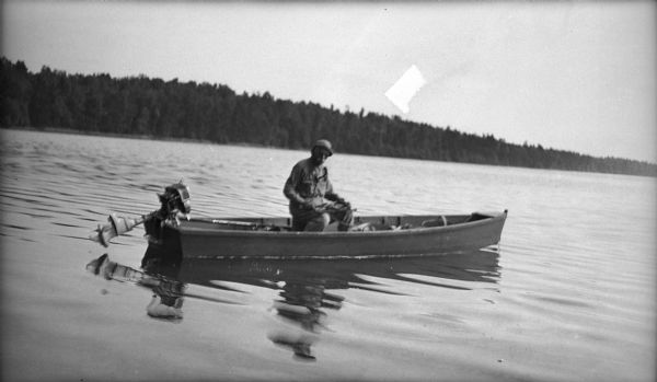 Dr. Birge sitting in a boat, probably at Trout Lake. The outboard motor on the boat has been tilted out of the water. A wooded shoreline is in the distance.