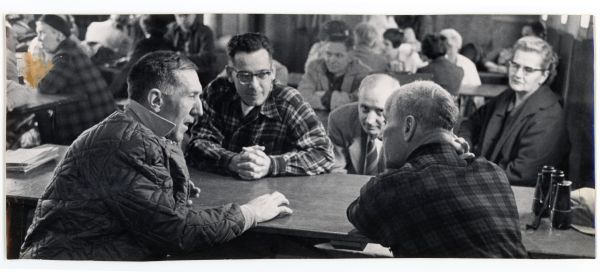 Four men and a woman are sitting at a table having an informal discussion. There are binoculars on the table.