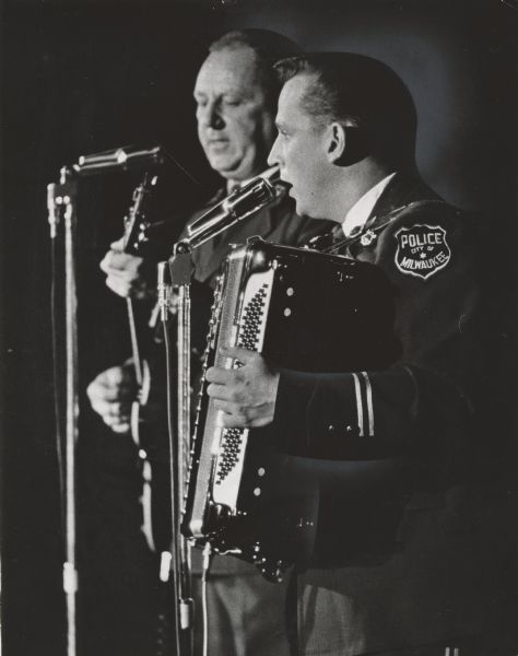 Two uniformed police officers stand in front of microphones, one holding a guitar and one holding an accordion. One officer's shoulder has a badge that says "Police, City of Milwaukee." Caption reads: <b>MUSICAL MEN IN BLUE</b> - The Milwaukee police department band held a concert and dance Wednesday in the Auditorium. Included in the 90 minute concert were numbers by Sgt. Arthur Rinderle on guitar and Patrolman Ronald Kleczka, accordion. Proceeds will be used to support the band, which receives no money from the police budget.