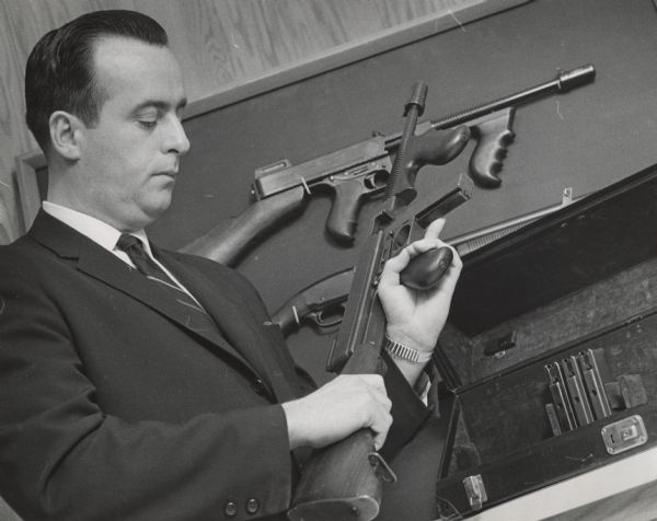 A man in a suit is holding and looking at a Thompson submachine gun. Another Thompson and a pump-action shotgun are hanging on the wall. Caption reads: "AN AGENT INSPECTS ONE OF THE GUNS STORED IN A VAULT IN THE FEDERAL BUILDING."