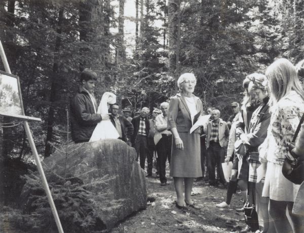 Derek Olson, standing on left unveiling stone, and Johanne Uhrenholdt Johnson speaking outdoors to a group of people at the rededication ceremony.