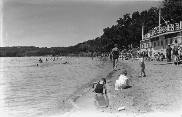 Several people in bathing suits and casual wear are relaxing on a beach and in the water at a Geneva Lake beach. Several people are also sitting behind a railing on the rooftop of a building at right. The sign on the building reads: "Go To Lakeview Inn For Chicken Dinner." The shore is tree-lined.
