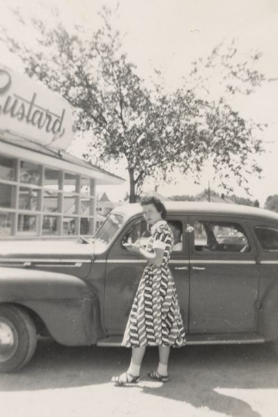A woman, perhaps a carhop, is standing outdoors by the passenger side of an automobile which is parked in front of Crawford's Custard. She is wearing a three-quarter length printed dress, and sandals. There is a man sitting in the driver's seat.