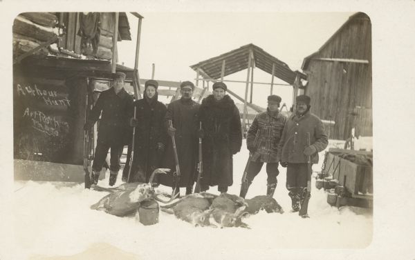 Handwritten on front: "A 4 hour Hunt." Handwritten on back: "4th man from left believed to be Teynor or Rural PD-C," and "An Art Roth Photo P.D.-C." The men are standing in the snow and are wearing coats and hats, and holding rifles. There is a log cabin on the left, and in the background is an open sided shed, and a barn on the right.