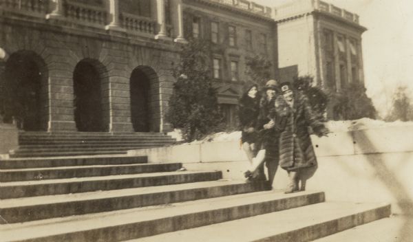 Three women wearing fur coats are posing alongside the steps leading up to the front of Bascom Hall.