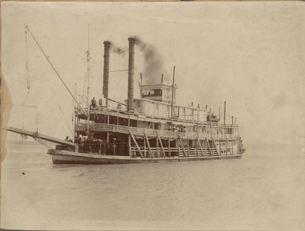 View across water towards the front left side of the Steamboat <i>Annie Laurie</i>. There is a man inside the pilot house, and the roof bell has what appears to be antlers mounted on top of it. A sign on the rail nearby reads: "U.S. Mail." Many of the male passengers are posing along the rail on the main and boiler decks of the steamboat. On the left at the bow is the stage suspended by a boom.