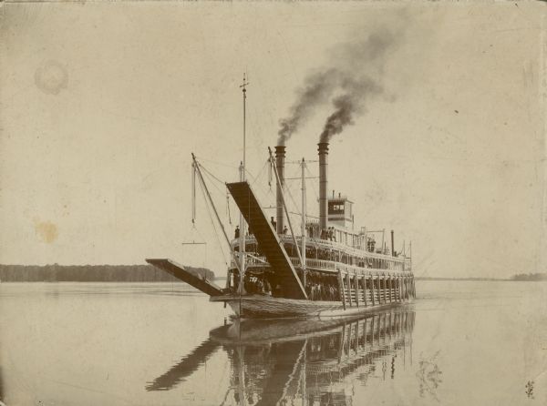 View across water towards the front of the <i>Saint Joseph</i>. The stages are suspended by the bows. Many of the passengers are posing along the front rails on the main and boiler decks of the steamboat. A wooded shoreline is in the distance.