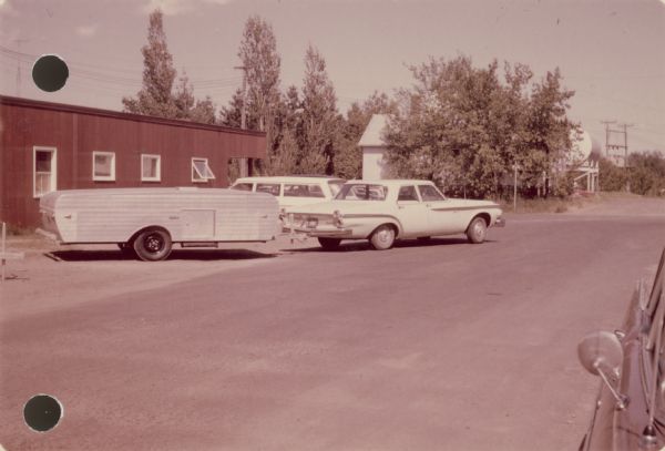 View down road towards an automobile pulling a folding trailer. Handwritten on back: "One of our trailers 'Rolite 16' at Grantsburg Wisconsin factory."
