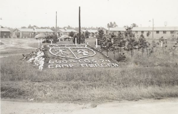 Camp Mercer sign spelled out with stones on a small, built-up hill. Camp buildings and parked cars are in the background.