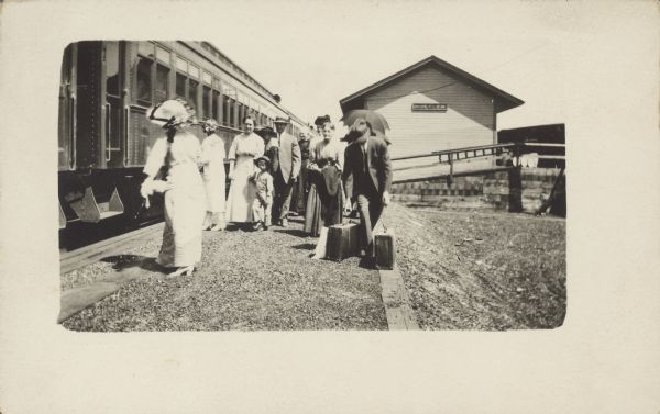Photographic postcard of men, women and children standing near a railroad train car at the Hustler Depot. Handwritten note along with postcard reads: "Reverse of picture has my grandmother's note: boarding trains after wedding for return to Montana. Bride Olga Nettlebeck, who is on the left with back to camera. Groom Samuel H. Crabtree getting the bags. Olga's mother Louisa Schroeder Nettlebeck — next to groom facing the camera."