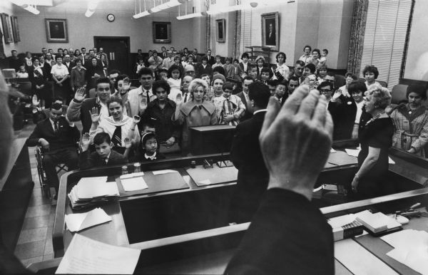 Several people are standing in a room, raising their right hands. In the foreground a person's hand is raised. Other people are standing in the background. Caption reads: "Twenty-three new United States citizens were sworn in Wednesday at the Federal building."