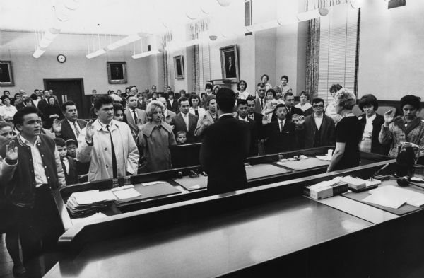 Several people are standing with their right hands raised. A man and a woman are standing and facing them. Caption reads: "<b>In time to celebrate Thanksgiving</b> as American citizens, 23 children and young people and two adults took the oath of United States citizenship Wednesday in the federal building. The oath was administered by Federal Judge Kenneth P. Grubb. It was one of the largest groups to receive the oath at one time, according to Morris Lieberman, attorney for the immigration and naturalization department."